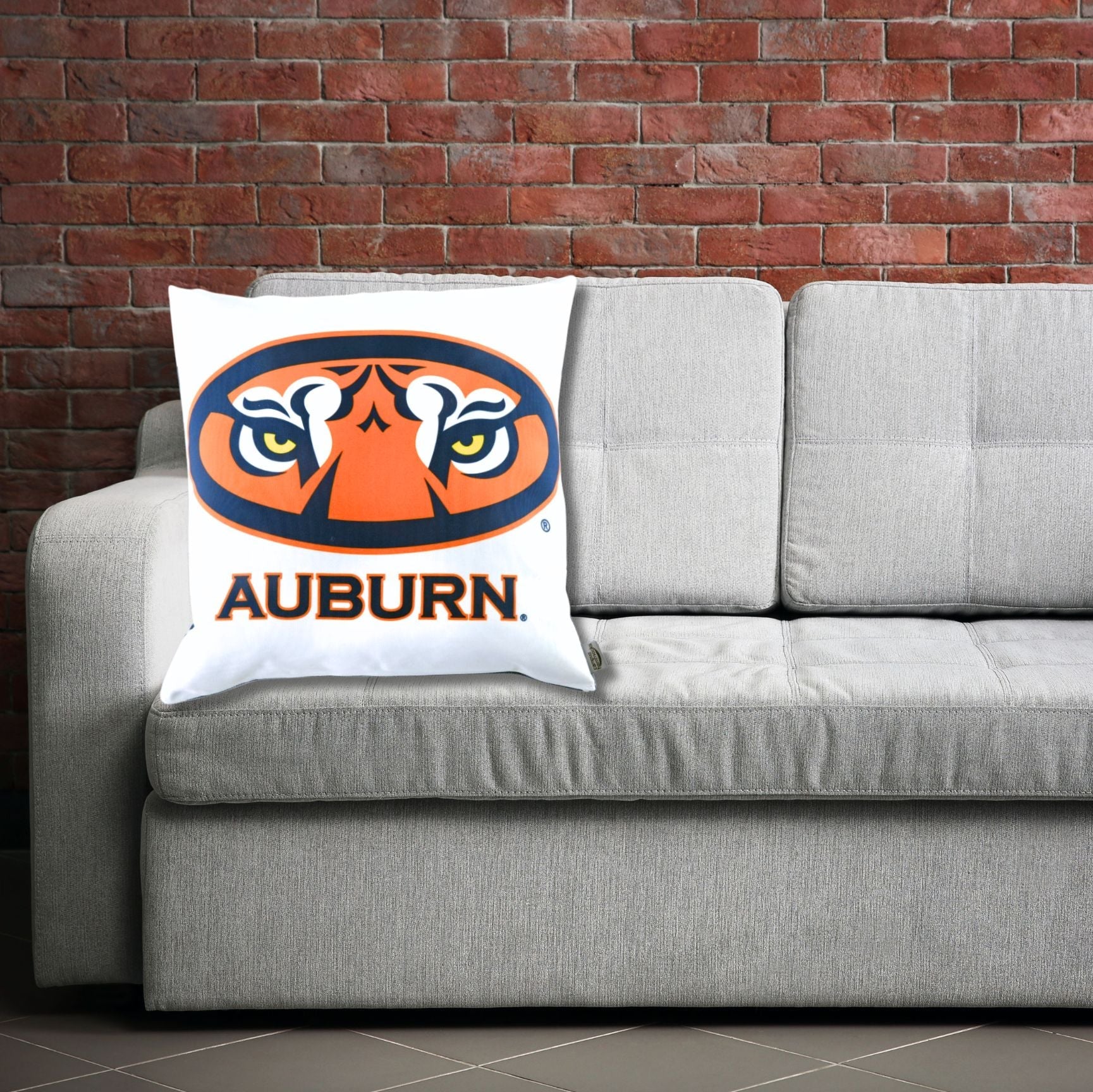 Gray sofa with an Auburn Tigers Eyes logo pillow against a brick wall.