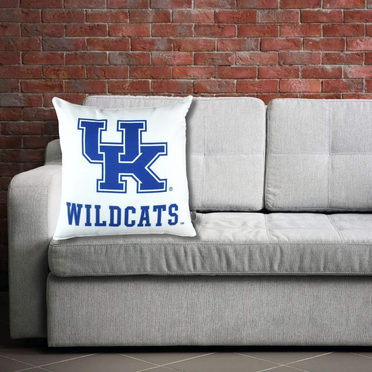 Gray sofa with a pillow featuring University of KEntucky Wildcats logo against a brick wall.