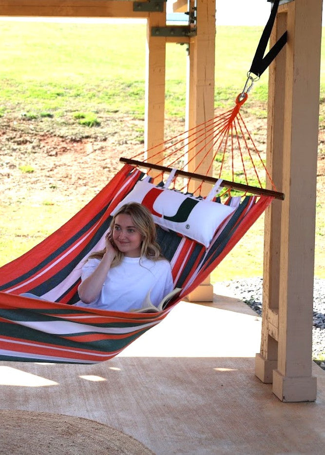 Person sitting in a colorful striped hammock under a wooden structure with a grassy field in the background.