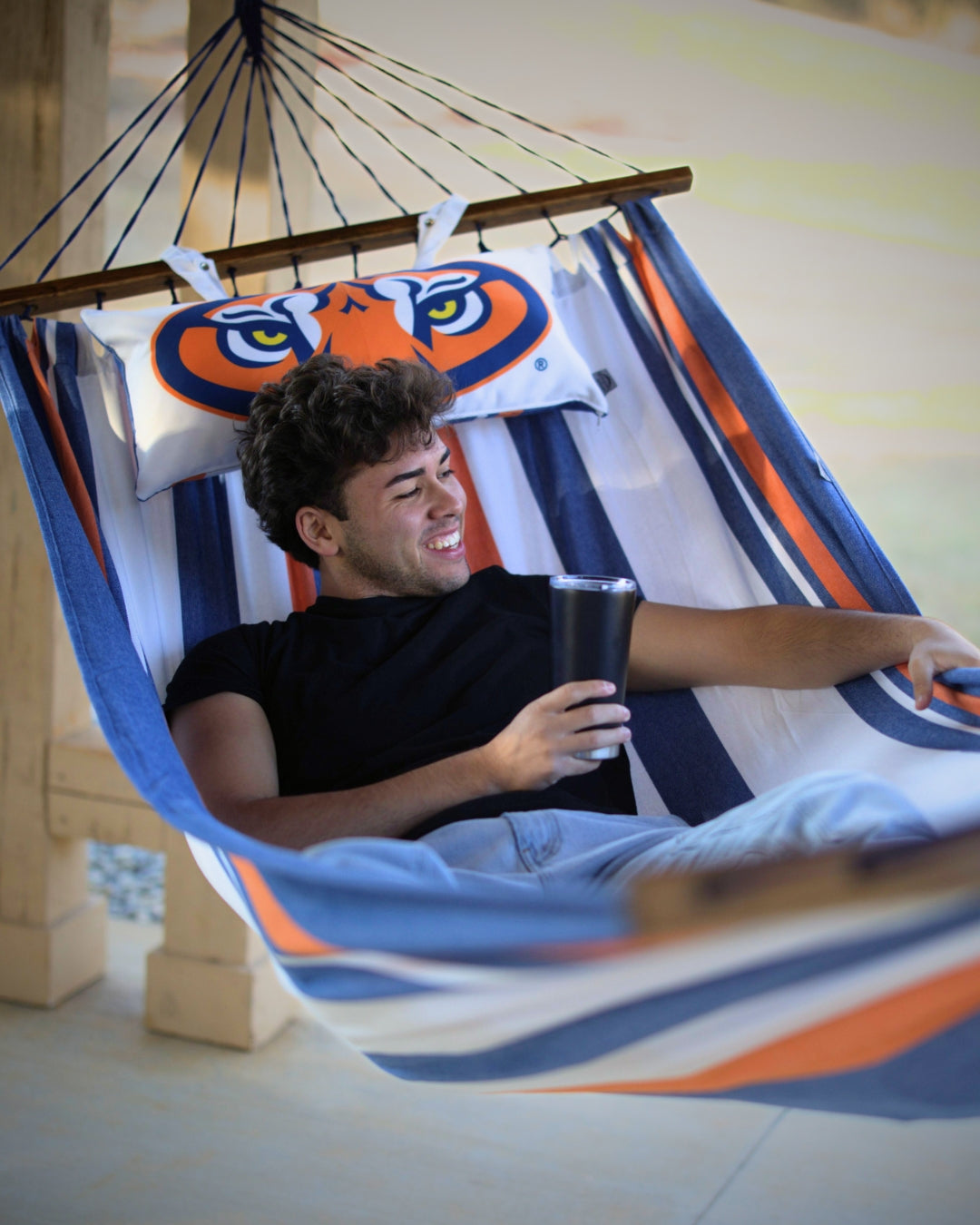 Man relaxing in a Auburn Tigers face logo hammock holding a cup, with a colorful pillow 