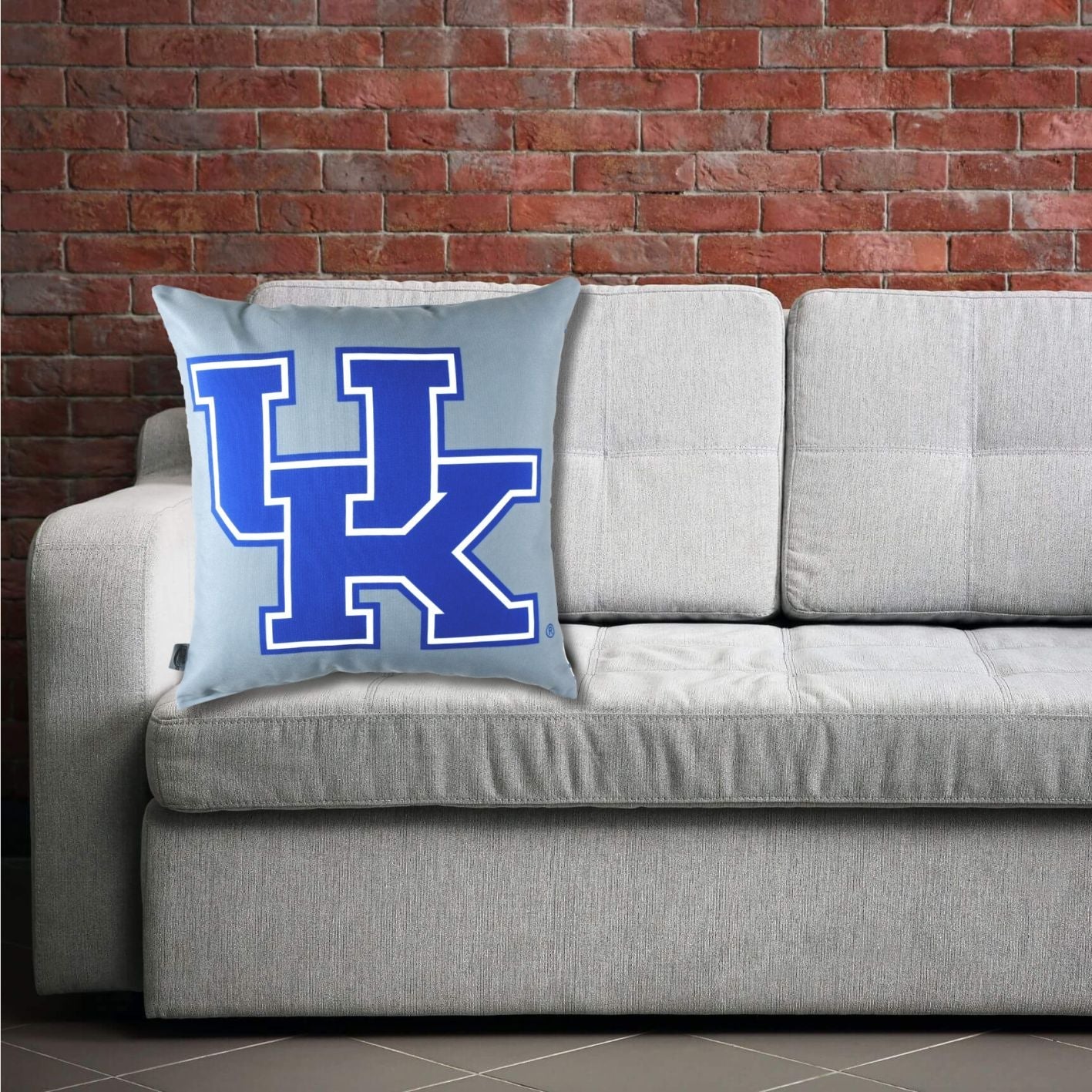 Gray sofa with a pillow featuring the UK University of Kentucky logo against a brick wall.