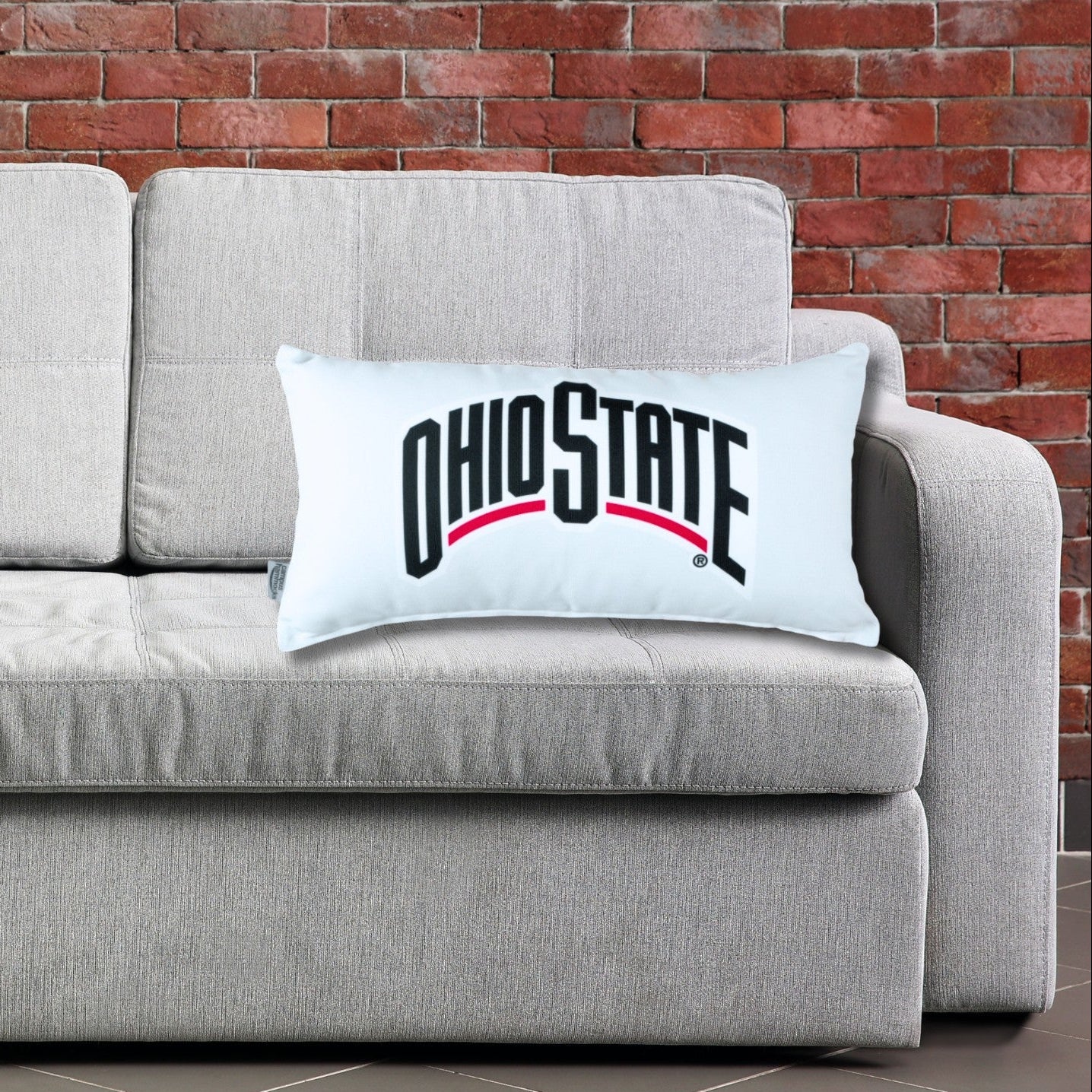 Ohio State-themed design throw pillow on a gray couch against a brick wall. Dorm room decor.